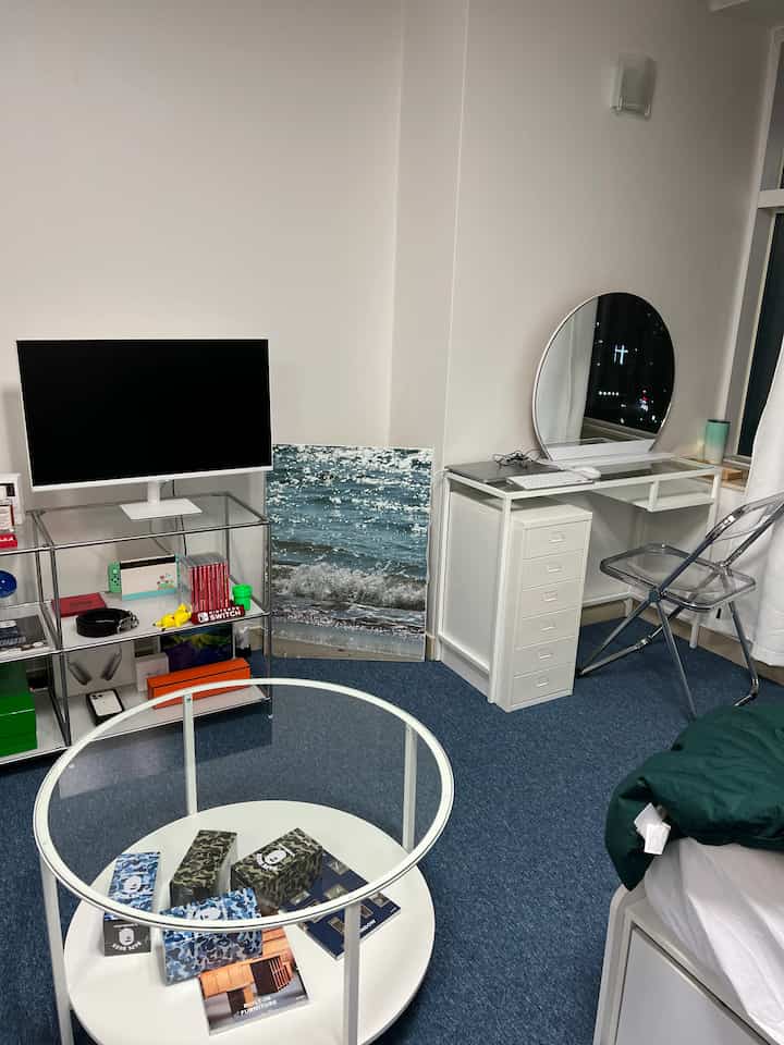 A modern small bedroom with white walls and blue carpet featuring a transparent chair and a vanity desk with round mirror
