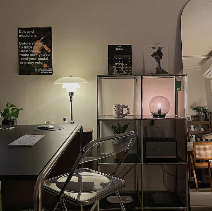 白い壁と黒いデスクが特徴のモダンなワンルームのホームオフィス、透明な椅子とモジュラースタンドの上に照明が配置された落ち着いた空間