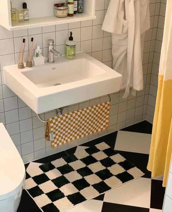 白黒のチェッカーボードタイルの床とディープグリーンのチェッカーボードバスマット、マスタード色のタオルと黄色のシャワーカーテンが配置されたバスルーム空間