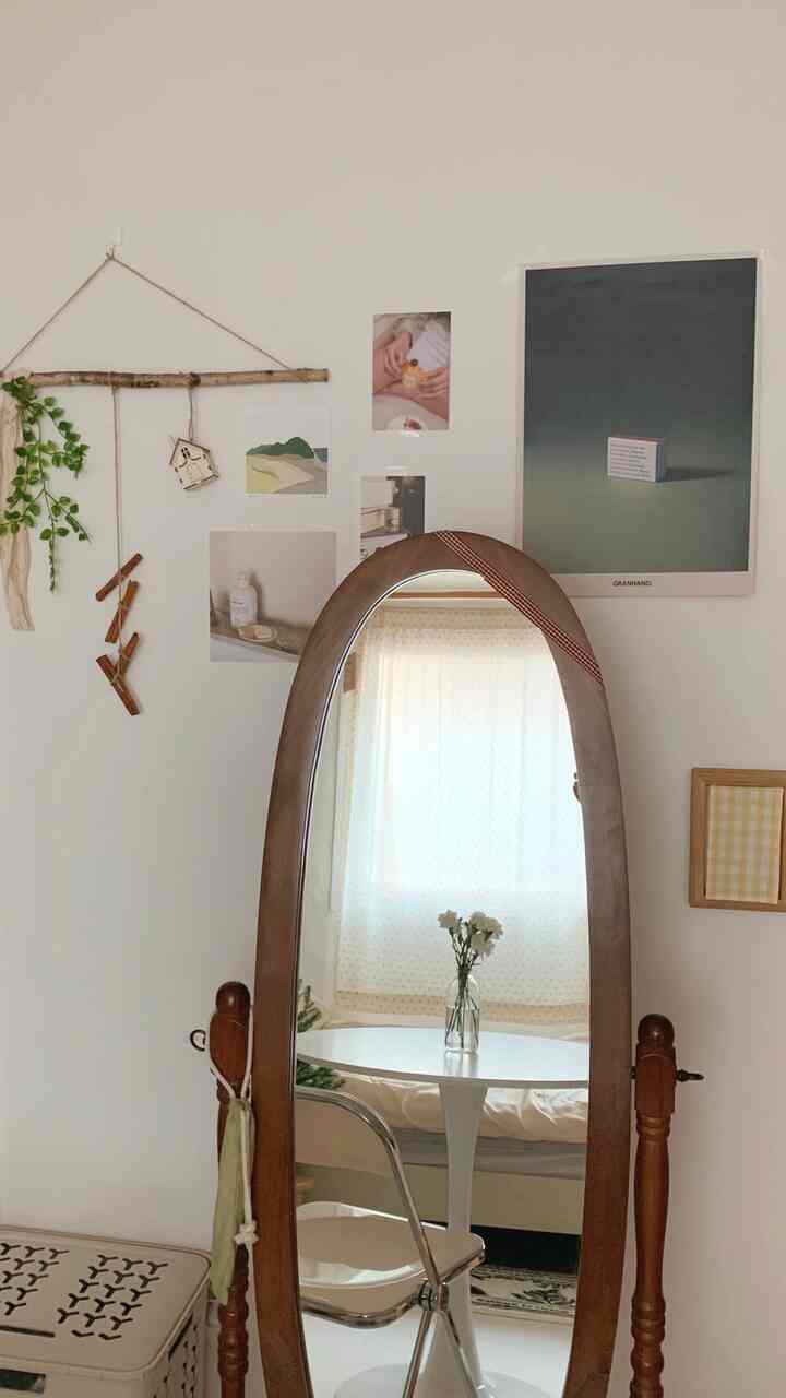 ナチュラルな色調のワンルーム、木製フレームの鏡とポスター、小さなダイニングテーブルに花瓶が置かれた雰囲気のあるインテリア