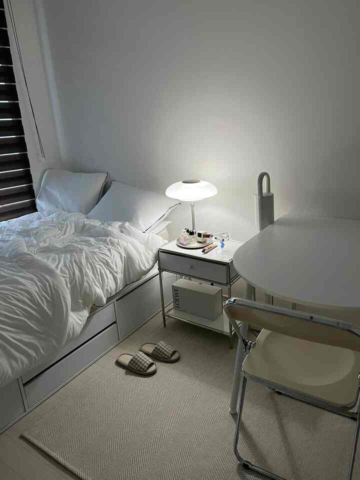 Simple studio bedroom in white and beige tones featuring a bed, nightstand, and table lamp with cozy atmosphere