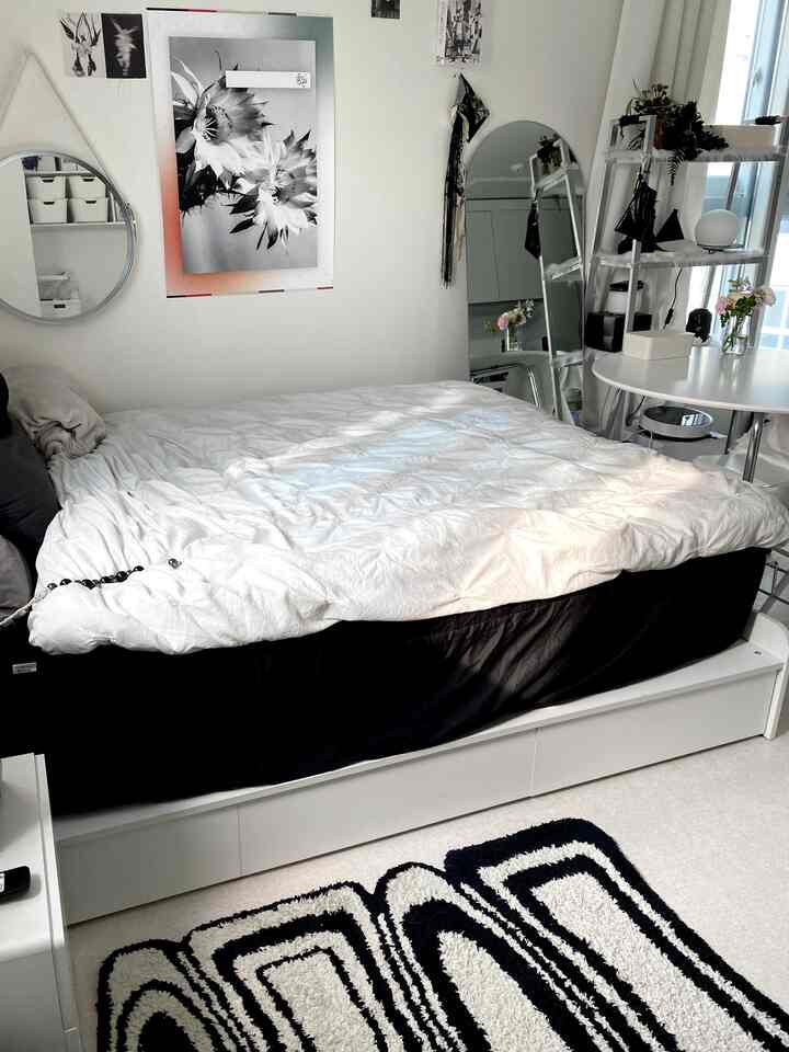 A modern bedroom with white and black tones featuring a centrally placed bed with white bedding, shelves on the right, and natural light blending harmoniously.