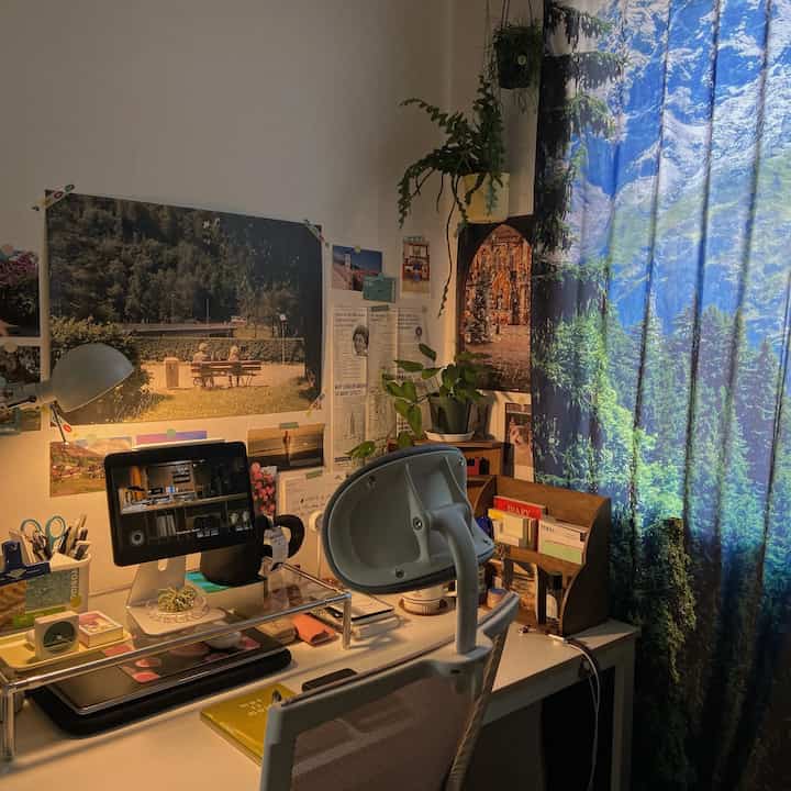 A cozy home office featuring a white desk and office chair, blue forest-themed curtain, and posters on the wall