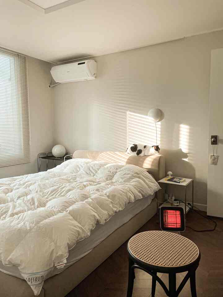Cozy bedroom in beige and white tones, featuring a cat sunbathing on the bed headboard and a space heater near the bed
