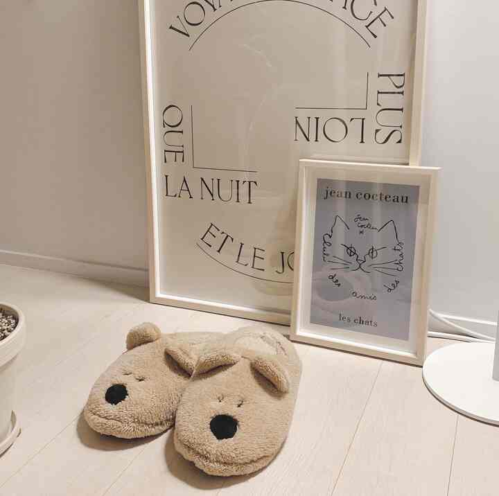 ベージュトーンの床と壁に2つの額縁が置かれ、その前に動物型のベージュ色スリッパがある暖かみのある空間