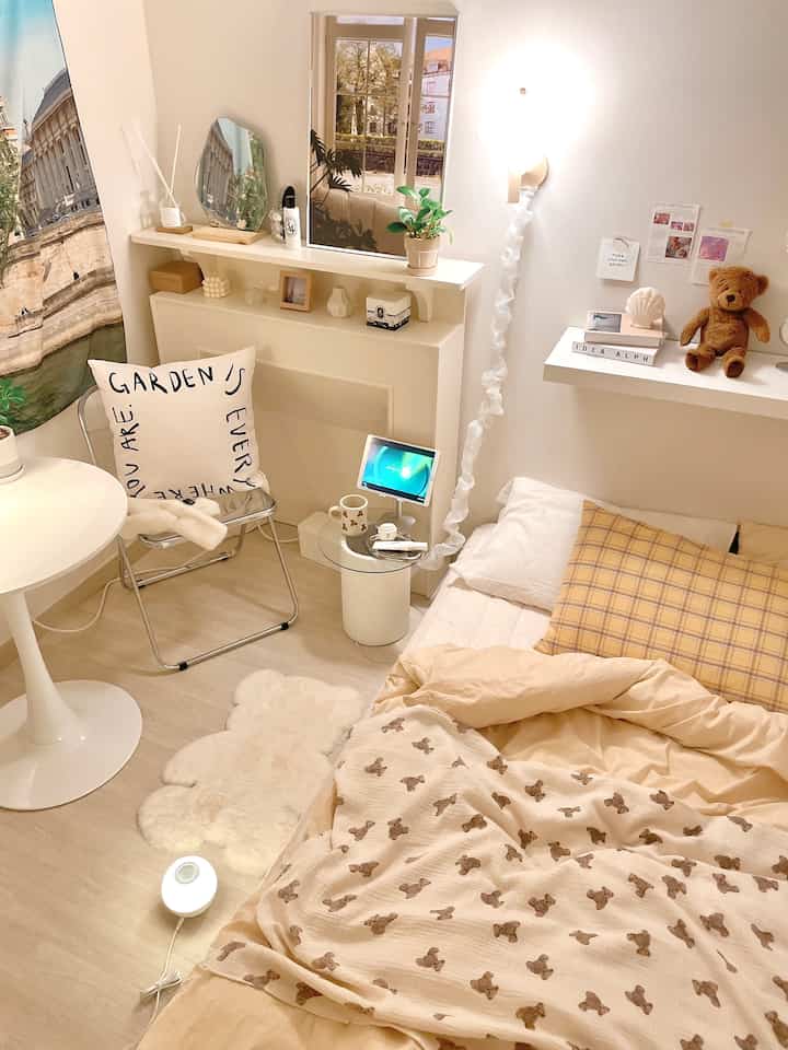 Beige and white toned bedroom featuring a transparent chair, round table, teddy bear patterned bedding, and wall shelves with cozy vibes
