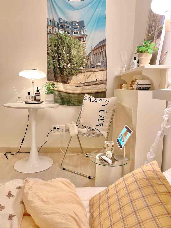 Cozy bedroom space in white and beige tones featuring a round table, transparent folding chair, and fabric poster with a simple decor