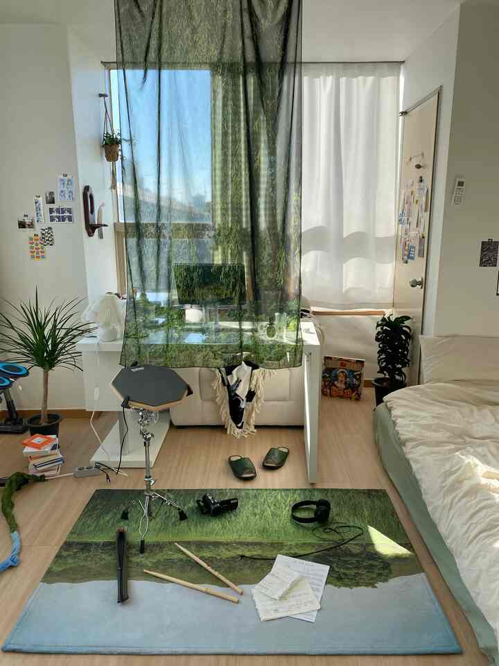 ナチュラルな色調のワンルームホームオフィス、デスク、ベッド、ラグ、植物が配置されたシンプルな雰囲気のインテリア