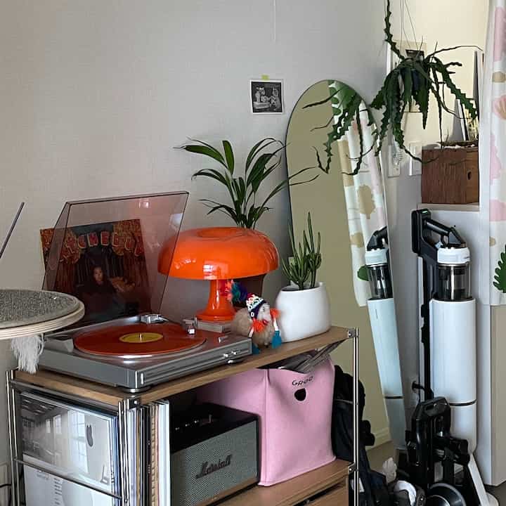 Studio apartment space with white walls and wood-tone stainless steel shelving, featuring an orange lamp, turntable, and plants in a tidy interior