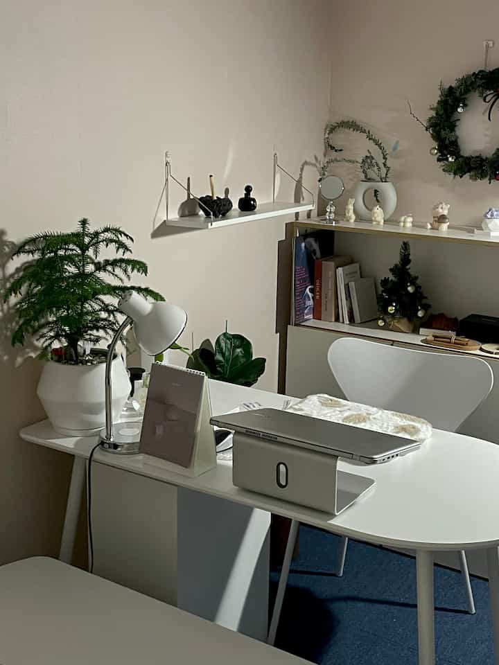 White-toned study featuring a central laptop on a semi-oval dining table, wall shelf, and dresser in a modern atmosphere