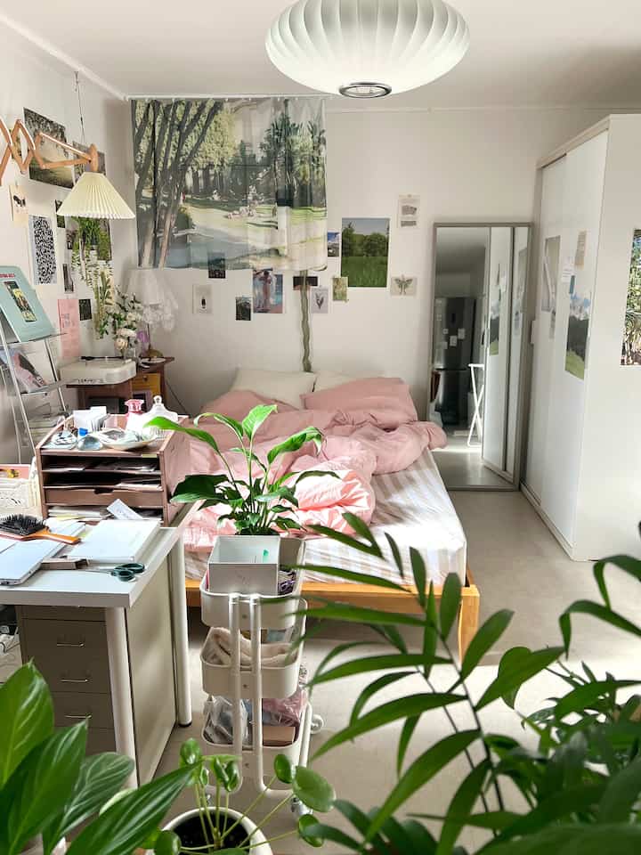 Natural-toned studio bedroom featuring pink bedding, lush plants, and simple furniture creating a cozy atmosphere
