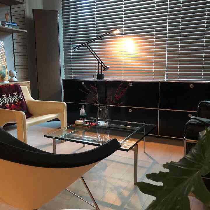 Black and white toned home office space featuring a tempered glass table, USM cabinet, modern armchairs, and blinds with cozy atmosphere