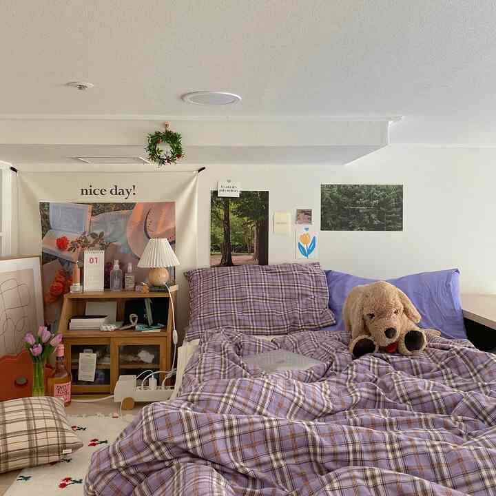 Studio apartment bedroom with white walls and ceiling, purple check duvet cover, cozy stuffed dog plush, and wooden nightstand creating a warm atmosphere
