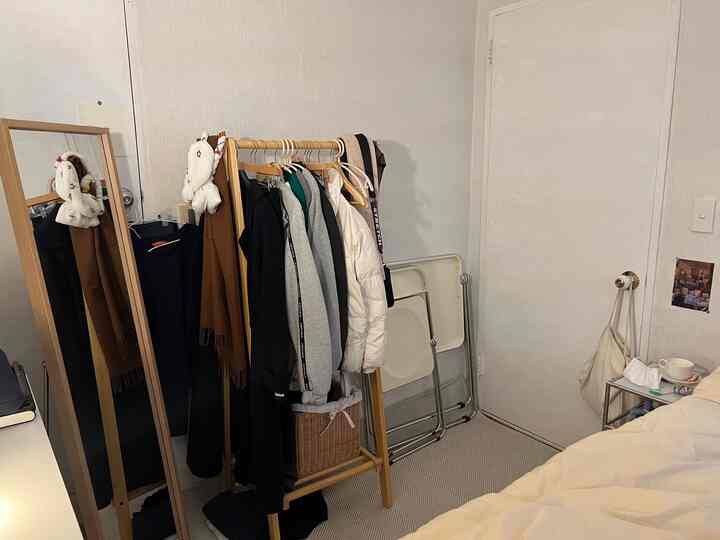 A compact bedroom featuring white and wood tones, with a clothes rack and mirror arranged for practical use