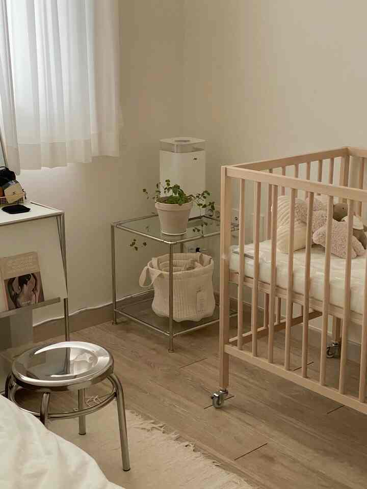 A simple bedroom with white and wood tones featuring a baby crib, stainless modular side table, and humidifier nearby