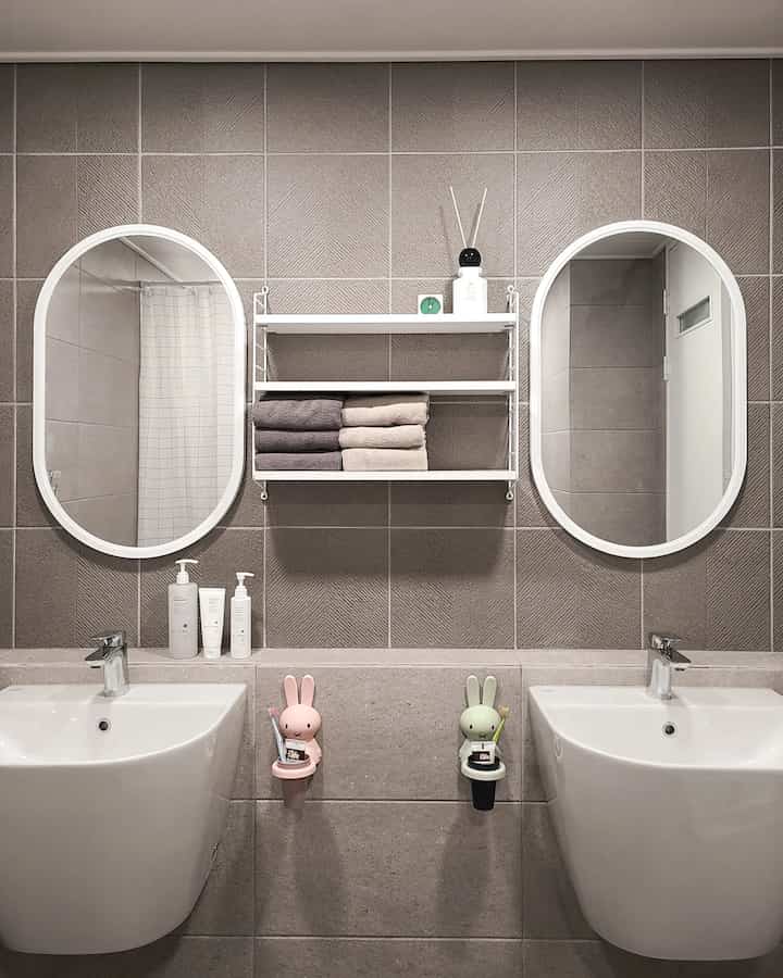 Gray and white toned bathroom featuring twin white sinks and mirrors with a central white shelf and pastel rabbit toothbrush holders creating a neat atmosphere
