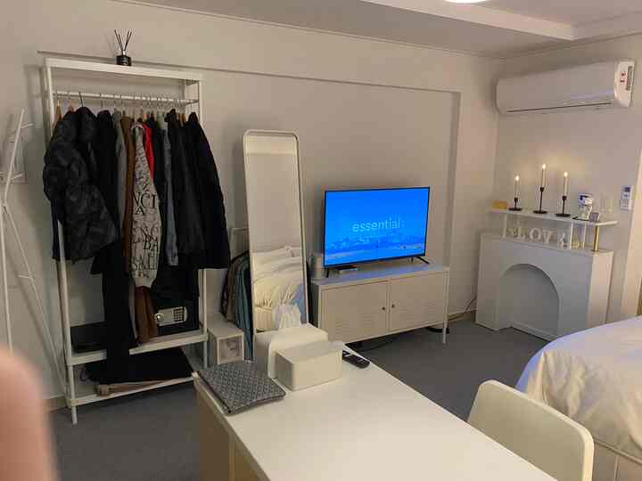 White and gray toned studio bedroom featuring a clothing rack, full-length mirror, TV cabinet, and desk in a modern, hotel-like interior