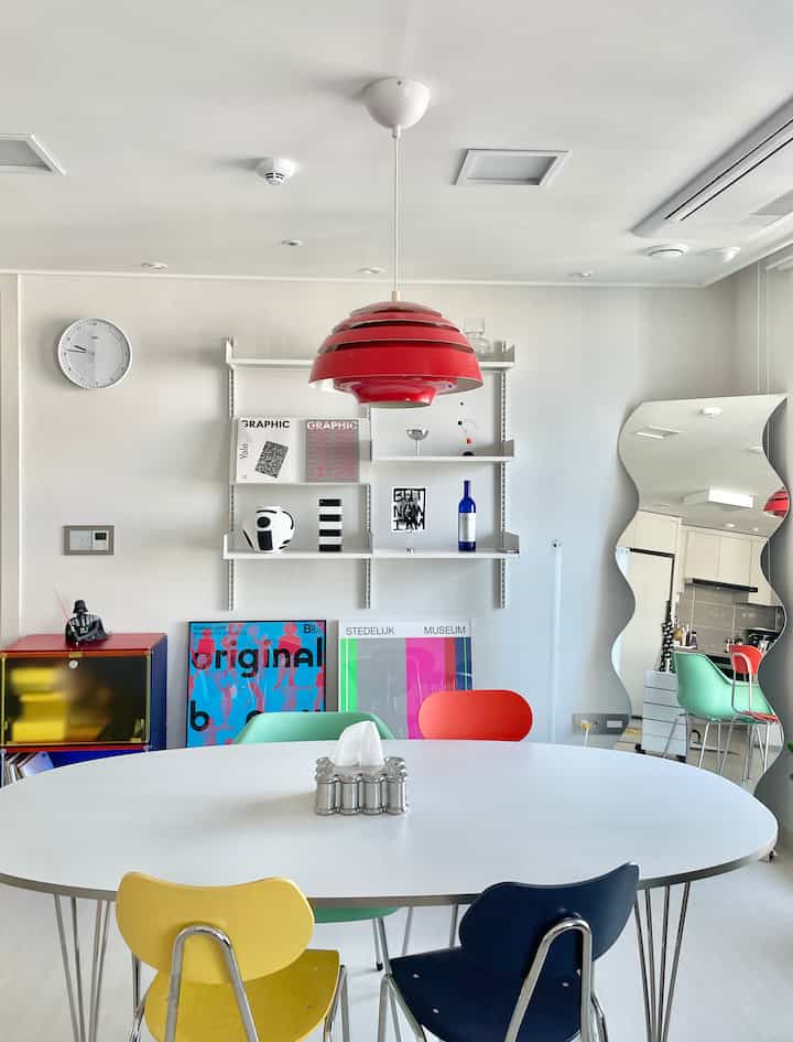 Bright white dining room featuring vivid colored chairs around a white oval table and a red pendant light suspended centrally