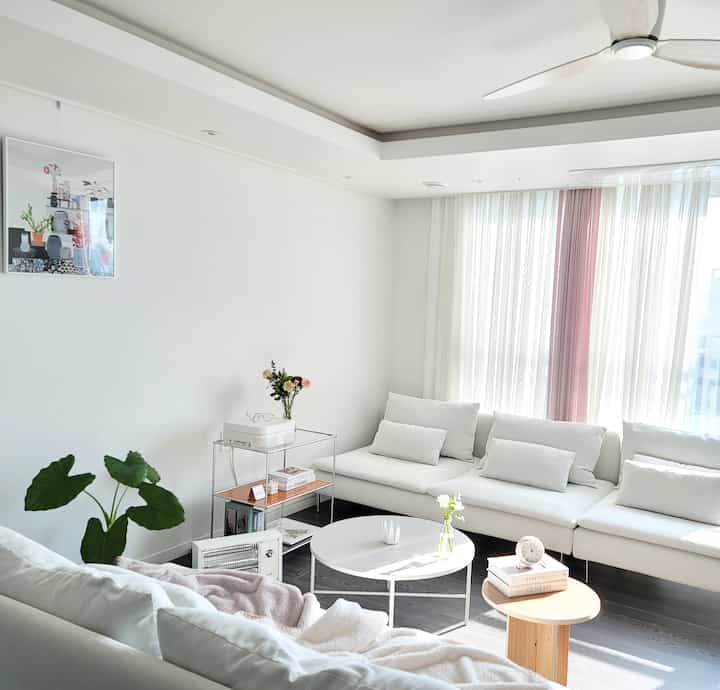 Bright, clean white-toned living room featuring large white sofa, sheer curtains, and wooden side table