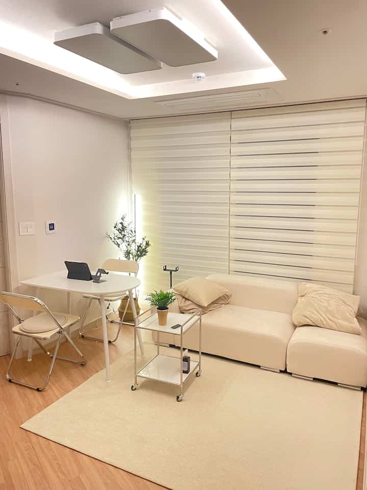Bright ivory and white-toned living room featuring an ivory sofa, white folding table, indirect lighting, and plants creating a warm cozy atmosphere