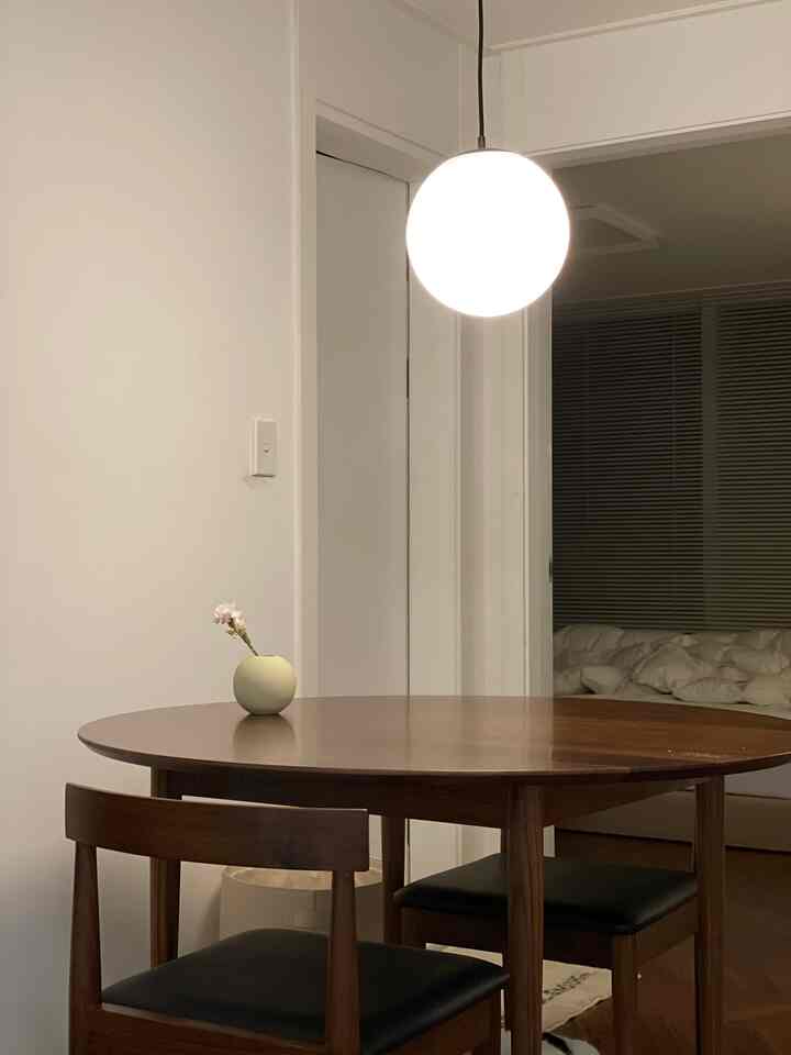 Simple two-person dining room with white walls and wood-tone furniture, featuring a round table with a small vase and a spherical pendant light