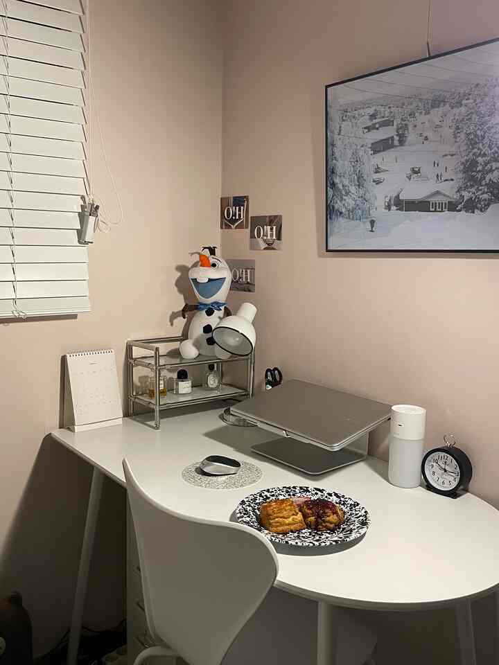 Beige-walled small home office featuring white furniture, laptop stand, and a plate of pastries evoking a cozy home cafe atmosphere