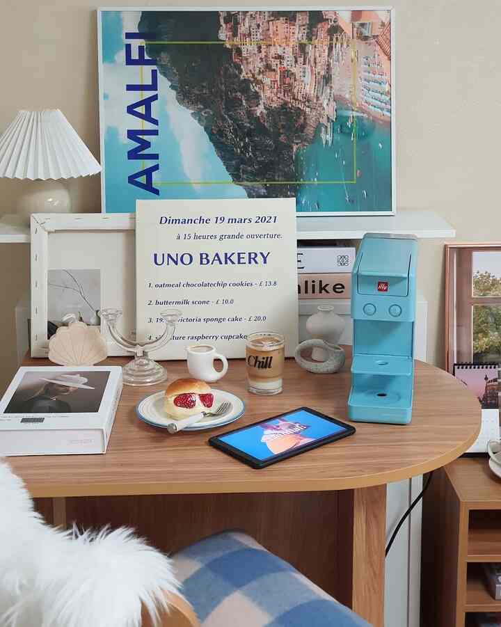 ウッド調のテーブルにブルーのコーヒーマシンとコーヒーカップ、ポスターや照明が配置された雰囲気のあるおうちカフェ空間