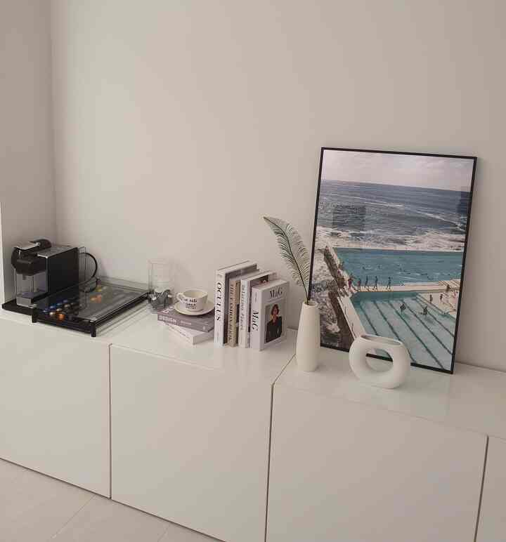 明るくミニマルなホワイトトーンのホームカフェ空間。コーヒーマシン、大型の額縁、白い花瓶がモダンに配置されたインテリア