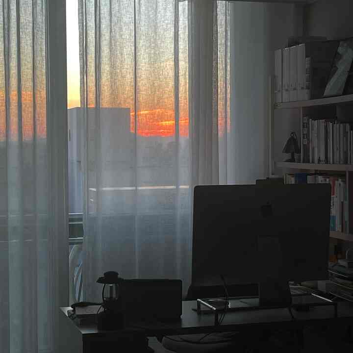 White sheer curtains reveal a sunset view in a living room home office with iMac and bookshelf creating a cozy workspace