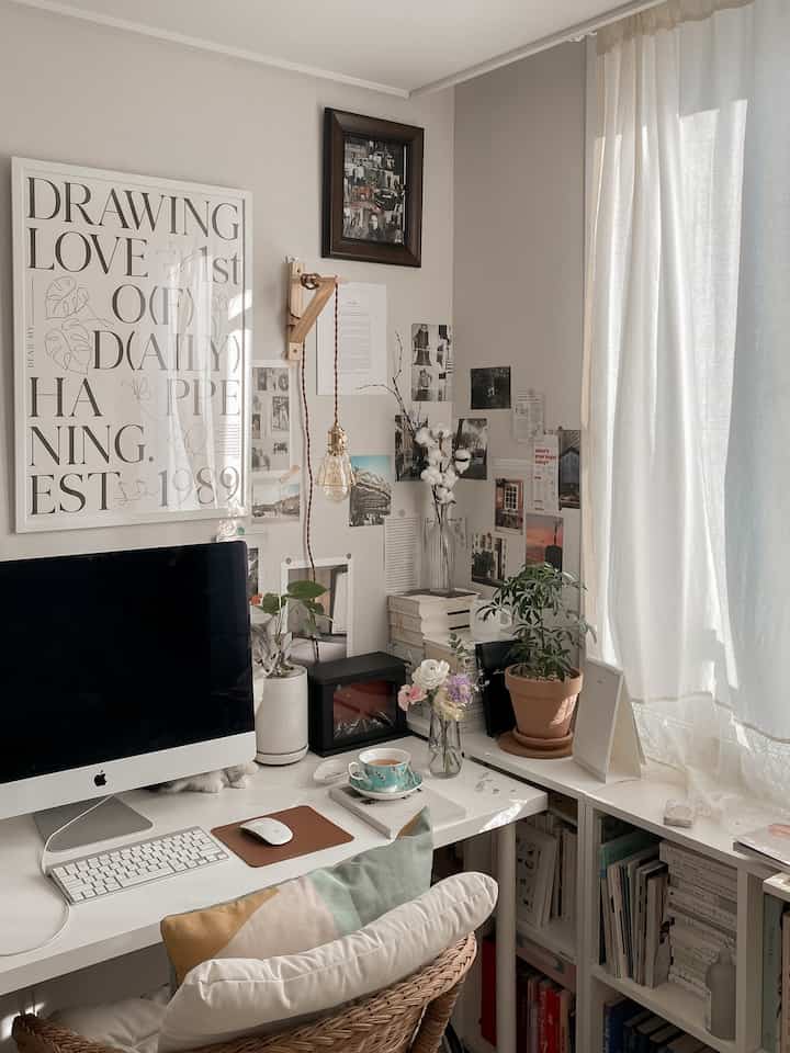 A white and natural tone home office featuring a desk, bookshelf, plants, and curtains creating a cozy atmosphere