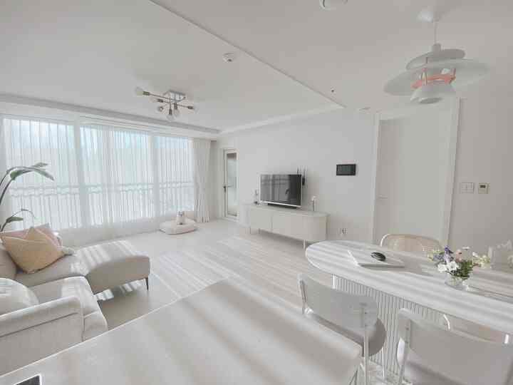 明るいホワイトトーンのリビングルーム、ライトベージュのソファとホワイトのダイニングチェアが配置されており、ペットベッドとカーテン越しに陽光が差し込むモダンなインテリア