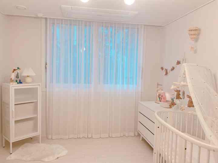 White-toned baby room featuring a round crib, sheer curtains, and rattan pendant light with a cozy natural atmosphere