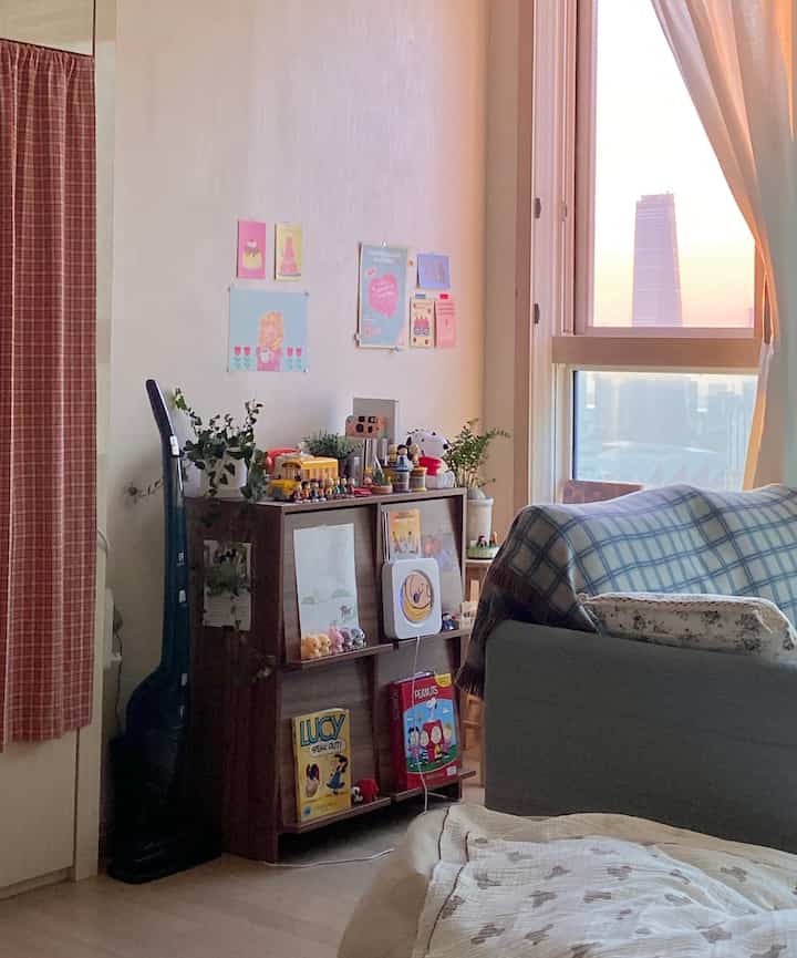 温かみのあるアイボリーと木目調のワンルームの窓辺、チェック柄ソファとポスター、植物が配置された居心地の良いインテリア