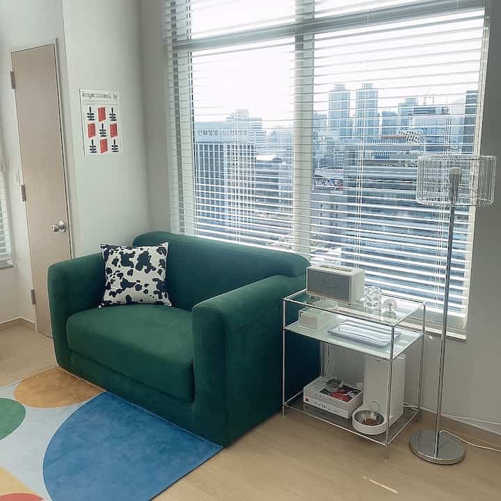 Modern living room with white walls and wood tone flooring, featuring a green sofa with patterned cushion, glass side table, and floor lamp in a clean space