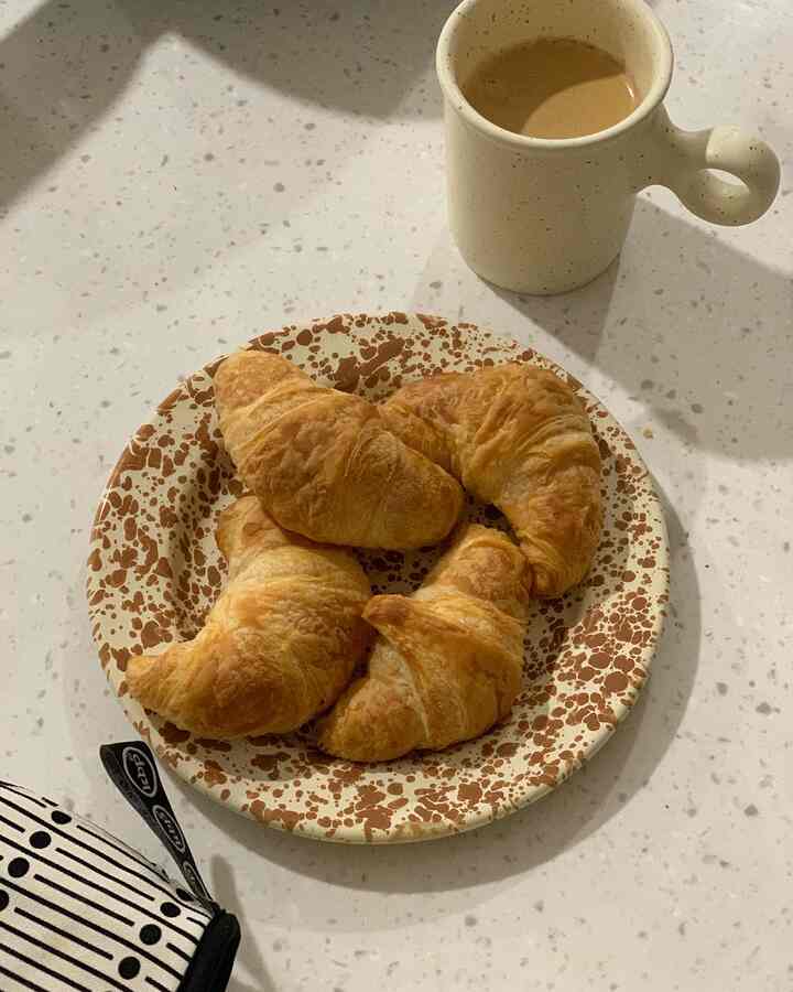 ベージュとナチュラルトーンのキッチン空間、茶色の模様がある皿にクロワッサンが置かれ、クリーム色のマグカップが一緒に配置された雰囲気のあるおうちカフェ