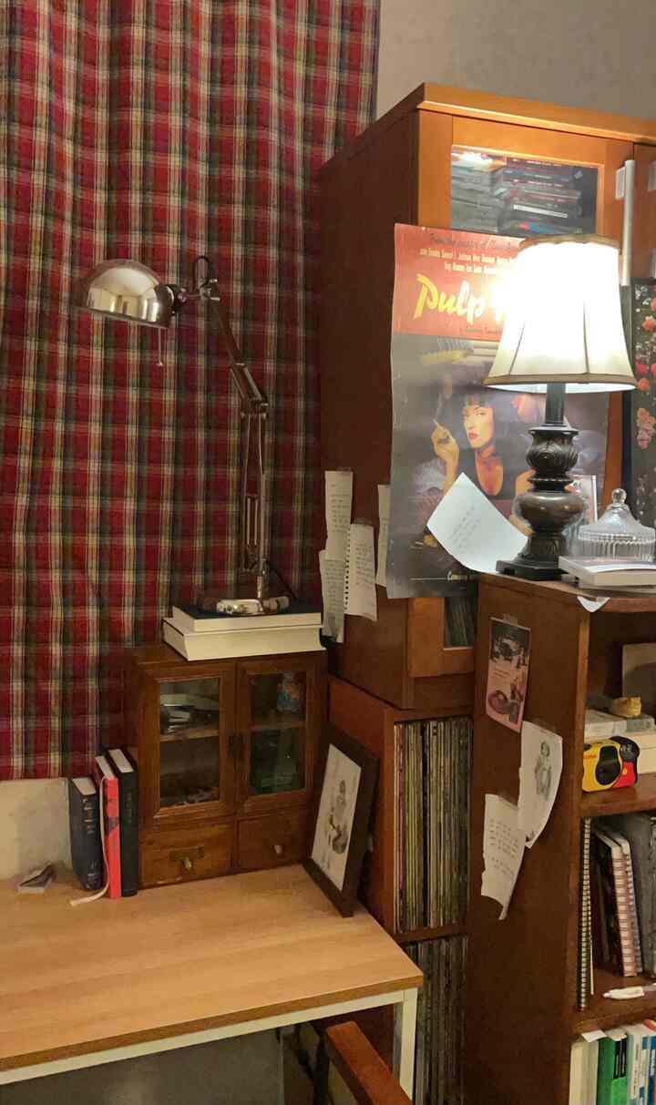 A vintage-style desk area with red and green plaid curtains as background, featuring a table lamp and shelving creating a cozy atmosphere