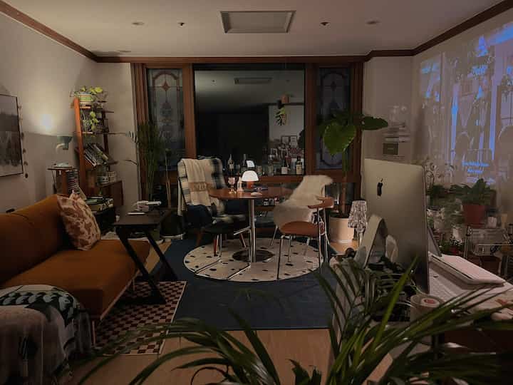 暖かいナチュラルカラーのリビング、円形のダイニングテーブルとソファ、多数の植物が配置された居心地の良い空間