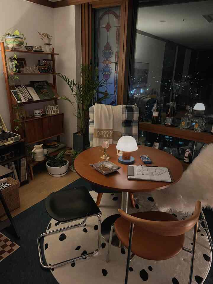Cozy cafe-style studio apartment featuring wooden furniture, dining table with chairs, plants, and a table lamp by the window with night city view