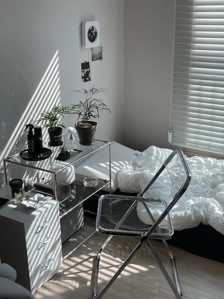 白と黒の落ち着いたトーンの寝室、透明な椅子と金属の棚、観葉植物が配置されたモダンで清潔感のある空間