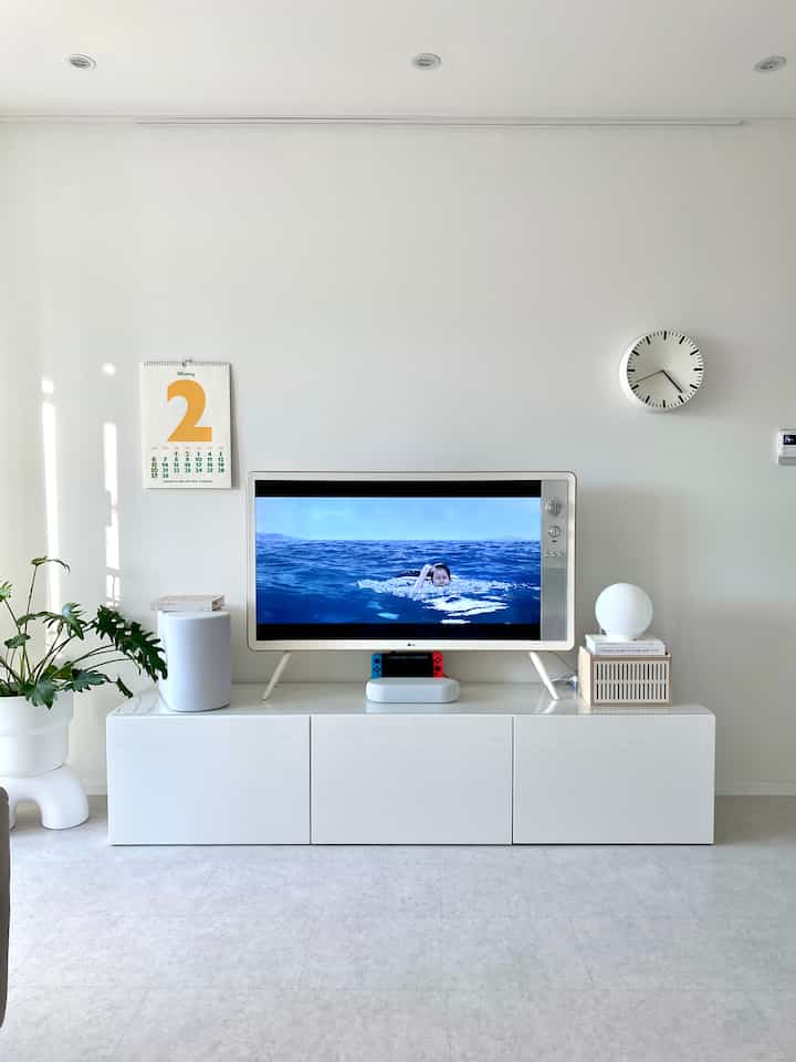ホワイトトーンの小さなアパートのリビング、中央にテレビとテレビ台、左側に植物、右側に時計が配置されたミニマルな空間