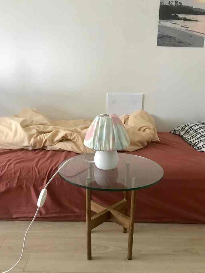 Studio apartment bedroom with white walls, beige duvet, brown bedspread, featuring a glass side table holding a pastel-tone lamp in a simple setting