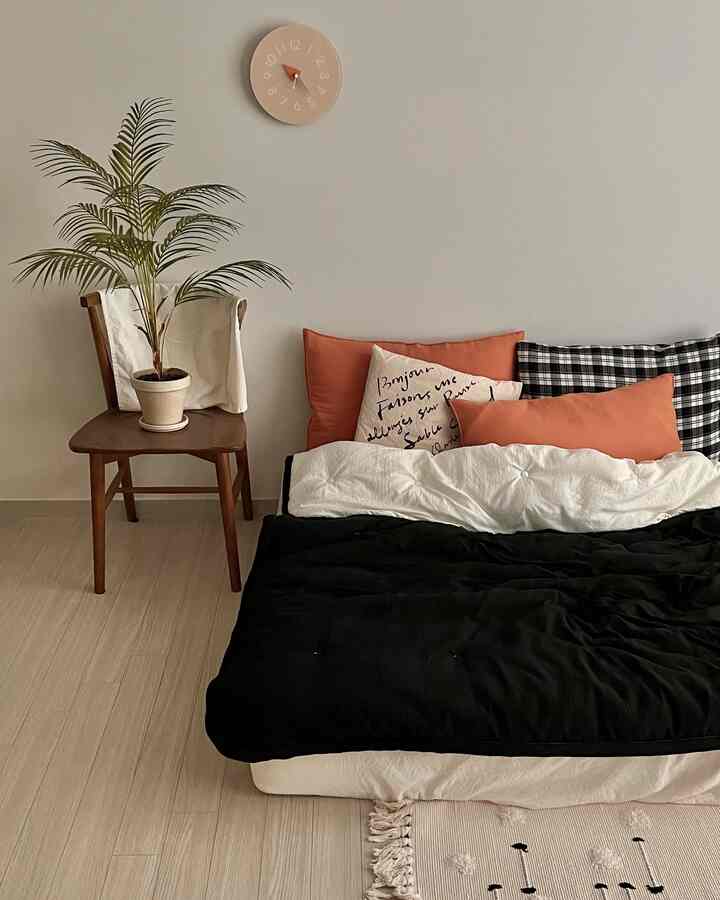 Natural toned bedroom featuring a bed with black comforter and orange cushions, wooden chair, and beige wall clock creating a cozy atmosphere
