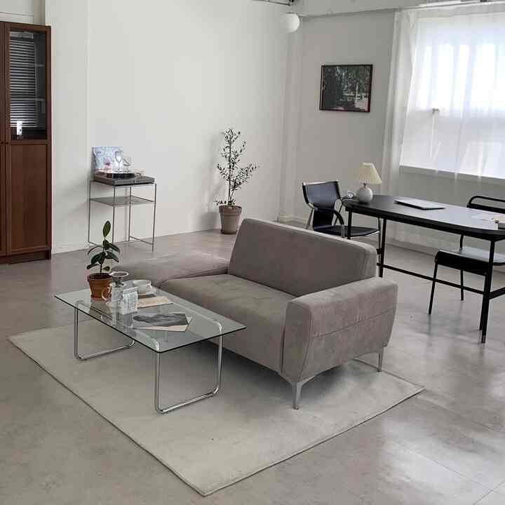 White and beige toned mid-century modern living room featuring a grey sofa, glass table, bookshelf, and plants in a clean photo studio space