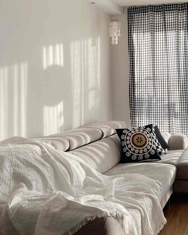 白とグレーのトーンのリビング、洗練されたコーナーソファとパターンのクッション、チェック柄カーテンが調和したナチュラルモダンな空間