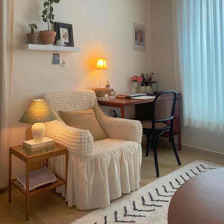 A cozy small living room-style home office in white and brown tones featuring a comfortable armchair, wooden desk, and natural plants