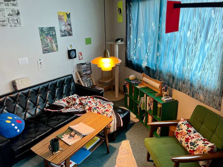 Cozy study with black leather sofa and green armchair, featuring yellow pendant light and a cat tower by the window