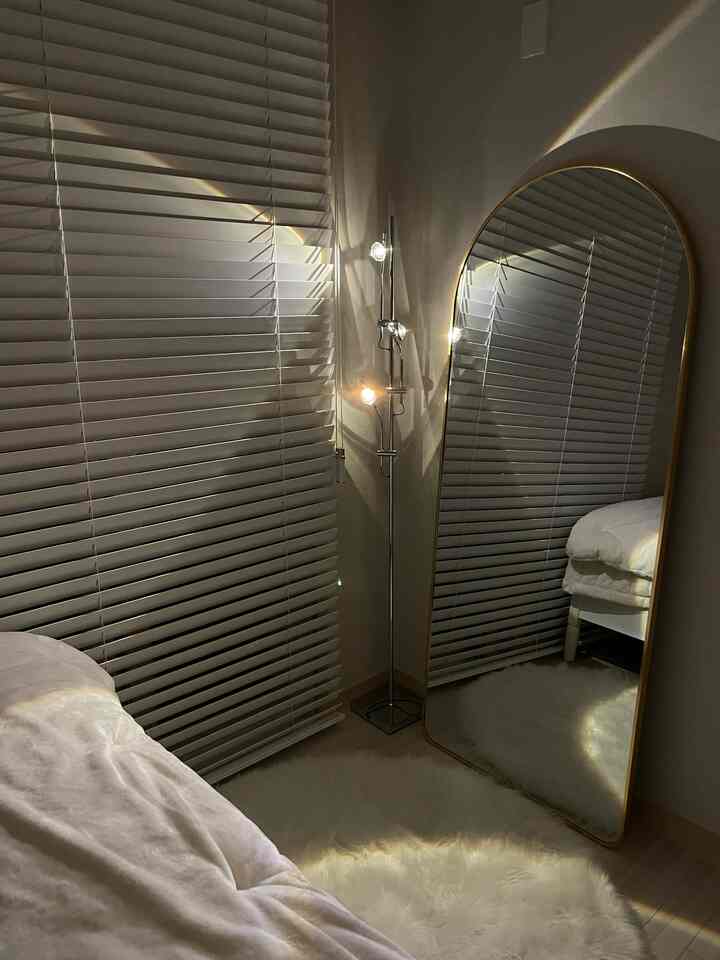 A white-toned bedroom corner featuring an arch-shaped gold framed full-length mirror, wooden blinds, mood floor lamp, and fluffy rug creating a cozy atmosphere