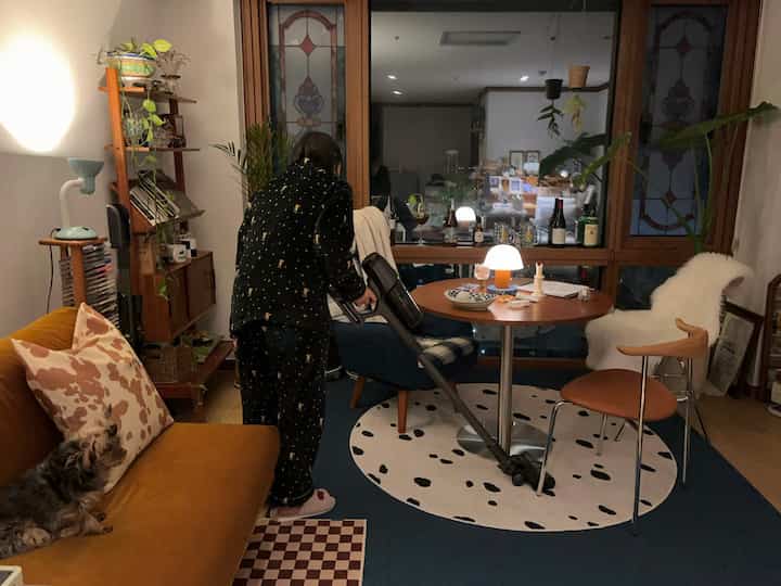 Warm natural-toned living room with a mustard sofa featuring a small dog, round dining table, and table lamp creating a cozy atmosphere