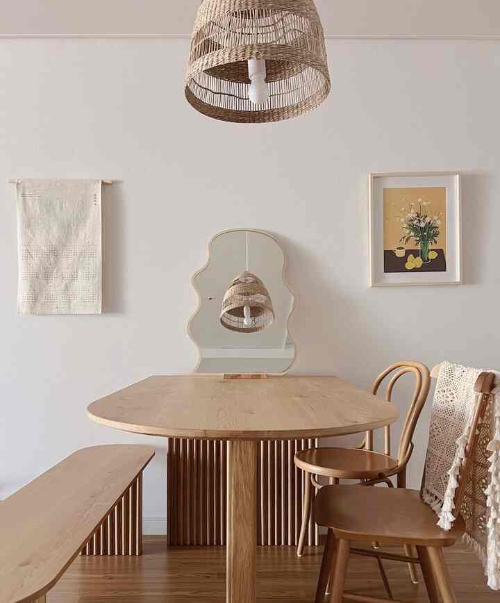 Beige and natural toned dining room featuring a wooden dining table, chairs, bench, rattan pendant light, and a uniquely shaped mirror creating a harmonious space