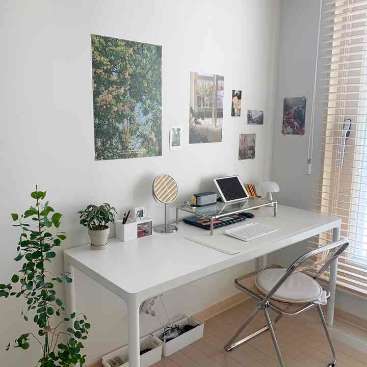 White and natural toned home office featuring a white desk, transparent chair, posters on wall, and green plants in a neat study setup